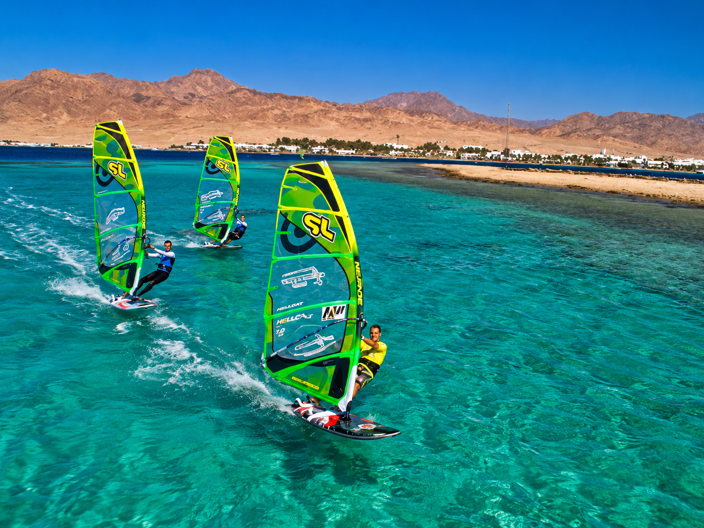 Three windsurfers racing across turquoise Red Sea water with desert mountains behind